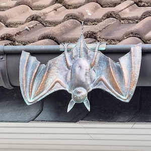 Pode incluir: Um ornamento de calha em forma de morcego de cor bronze. O morcego tem as asas abertas e é preso à calha com um clipe de metal. O corpo do morcego tem uma superfície texturizada e as asas têm veias detalhadas.