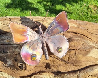 May include: A handcrafted metal butterfly sculpture with iridescent wings in shades of orange, pink, and purple. The butterfly has a detailed body and antennae, and is adorned with two yellow and green gemstone accents. The sculpture is displayed on a piece of weathered wood.