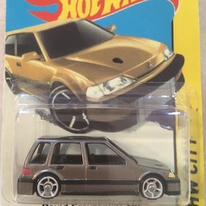 May include: A Hot Wheels die-cast toy car in a package. The car is a gold-coloured Honda Civic with black wheels. The packaging is blue and yellow with the Hot Wheels logo and images of race car drivers.