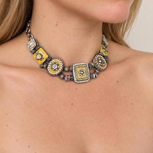 Op de afbeelding: Een choker-halsketting met zilverkleurige metalen schakels afgewisseld met gele geëmailleerde vierkante en ronde bedels, elk versierd met kleine heldere kristallen en gedetailleerde bloemmotieven langs de ketting.