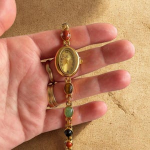 Peut inclure: Montre-bracelet dorée avec un cadran ovale et un bracelet orné de pierres précieuses colorées. Le cadran affiche les chiffres de 1 à 12. Le bracelet comporte une série de pierres précieuses rondes et ovales dans les tons de rouge, marron, vert et noir.