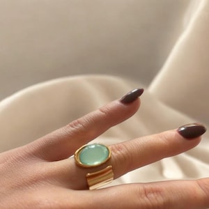 Op de afbeelding: Een goudkleurige ring met een lichtgroene ovale steen. De ring heeft een gespleten bandontwerp met een getextureerde, horizontale balk. De ring zit aan een vinger met donkere nagellak.