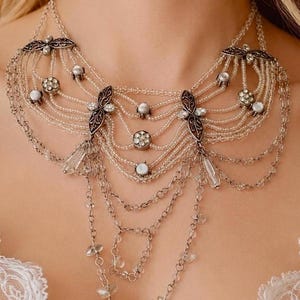 Op de afbeelding: Een meerlaagse statement ketting met heldere glaskralen, zilverkleurige metalen kettingen en gedetailleerde filigraan accenten. Het ontwerp bevat ronde strass-clusters en bungelende kristalachtige druppels voor een gedrapeerde uitstraling.