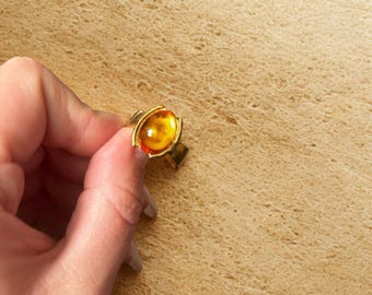 May include: A gold-toned ring featuring a large, oval-shaped amber stone. The amber has a warm, golden-orange hue, and the ring's setting has a simple, yet elegant design. The ring is held in a hand against a textured, tan background.