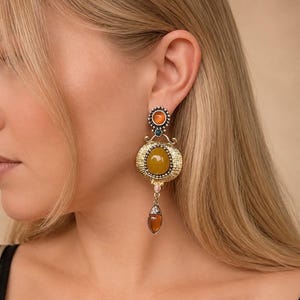 Op de afbeelding: Een goudkleurige hangende oorbel met een ronde oranje steen aan de bovenkant, een grotere ovale mosterdgele centrale steen in een getextureerd gouden frame en een kleine druppelvormige amberkleurige steen onderaan.