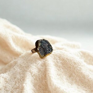 Op de afbeelding: Een unieke ring met een donkere, getextureerde steen met gouden accenten. De steen is gezet op een bronskleurige band. De ring wordt gepresenteerd op een crèmekleurige, getextureerde stof, wat een contrast van kleuren en texturen creëert.