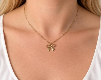 Puede incluir: Un collar de tono dorado que presenta una cadena delicada y un pequeño colgante centrado con forma de lazo. El metal tiene un acabado pulido y el colgante cuelga de una cadena de eslabones simples que descansa sobre la clavícula.