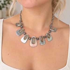 Op de afbeelding: Een zilverkleurige metalen ketting met een reeks hangende, schildvormige hangers. Elke hanger is voorzien van een gladde, ovale, doorschijnende bruingrijze steen in het midden, verbonden door kleine, gestructureerde ronde schakels.