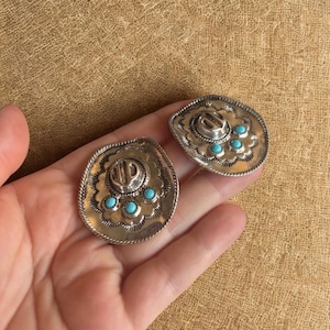 May include: A pair of silver-coloured earrings shaped like cowboy hats, each with turquoise-coloured stones. The earrings feature a detailed design with a rope-like border and a small hat shape in the centre. They are displayed on a tan surface.