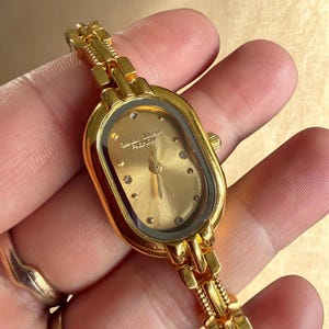 Può includere: Un orologio da polso dorato con quadrante ovale e cinturino a maglie. Il quadrante è dorato con piccoli accenti di diamanti. L'orologio è tenuto in mano, evidenziando il design dettagliato e la maestria dell'orologio.