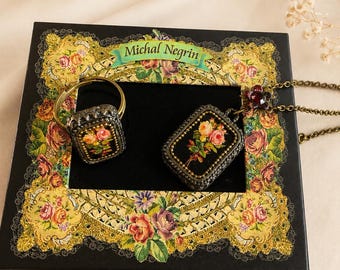Puede incluir: Un conjunto de joyería Michal Negrin que incluye un anillo rectangular y un collar con colgante a juego. Ambas piezas tienen una base negra con estampado de rosas, enmarcadas por un borde metálico con cuentas. Presentado sobre una caja negra floral.