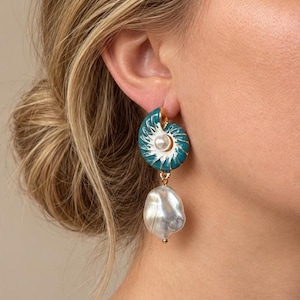 Op de afbeelding: Een hangende oorbel met een blauwgroene en witte nautilus-schelpvormige knop met een centrale witte parel, verbonden door een goudkleurige schakel aan een grote, onregelmatige, glanzende witte barokparel hanger.