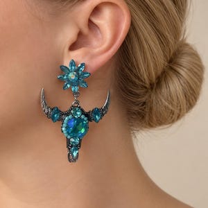Op de afbeelding: Een opvallende oorbel met een zilverkleurig stierenschedelmotief, versierd met felblauwe gefacetteerde kristallen en kleine turkooiskleurige kralen. Het bovenste deel is een bloemvormige cluster van blauwe kristallen.