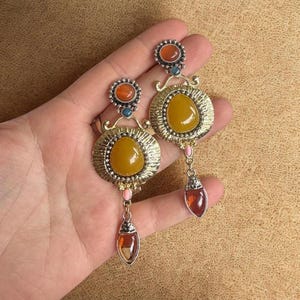 Op de afbeelding: Een paar sierlijke oorbellen met grote geel-oranje middenstenen, gezet in zilverkleurig metaal. De oorbellen hebben kleinere oranje en blauwe accentstenen, met een druppelvormige barnsteenkleurige steen die aan de onderkant bungelt.