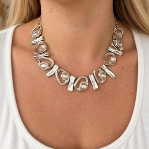 Op de afbeelding: Een zilverkleurige ketting bestaande uit een reeks onderling verbonden, organisch gevormde metalen schakels met een getextureerde, gehamerde afwerking. Elke schakel bevat een kleine, ronde, doorschijnende steen in het midden.