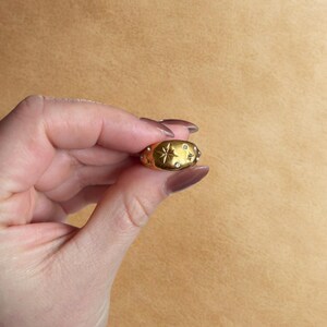 Op de afbeelding: Een goudkleurige ring met een sterrenpatroon en kleine, heldere edelstenen. De ring wordt in een hand gehouden, waarbij de details worden benadrukt. Het centrale ontwerp is een ster. De achtergrond is neutraal, wat het ontwerp en het vakmanschap benadrukt.