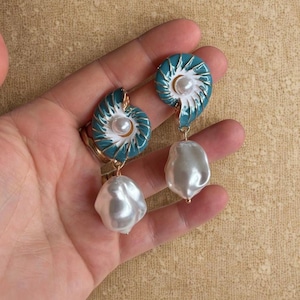 Op de afbeelding: Een paar oorbellen met een turquoise en wit schelpmotief met een parel in het midden. Elke oorbel heeft een grote, onregelmatig gevormde witte pareldruppel. De oorbellen worden in een hand gehouden.