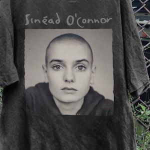 以下が含まれることがあります： シニード・オコナーの白黒ポートレートがプリントされた濃いグレーのTシャツ。歌手の名前は、ポートレートの上に明るいグレーの様式化されたフォントで印刷されています。Tシャツは木製のハンガーに掛けられています。