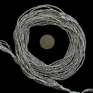 May include: A collection of silver-colored seed beads, strung together on white thread, arranged in a circular pattern. A U.S. quarter coin is included for size reference. The beads are small and have a metallic sheen, suitable for jewelry making or craft projects.