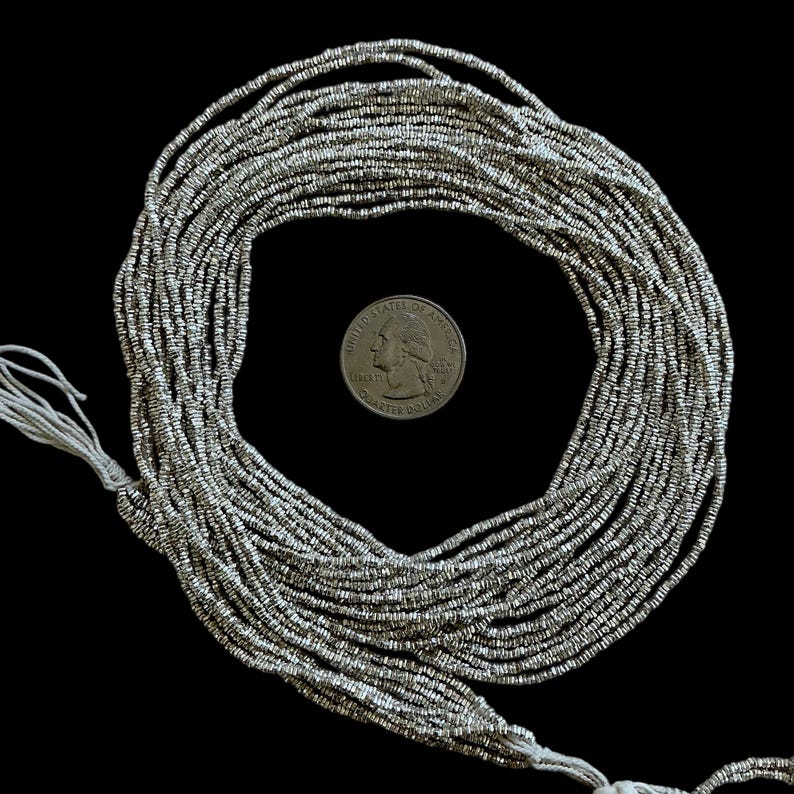May include: A collection of silver-colored seed beads, strung together on white thread, arranged in a circular pattern. A U.S. quarter coin is included for size reference. The beads are small and have a metallic sheen, suitable for jewelry making or craft projects.