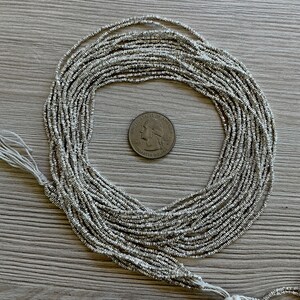 May include: Silver seed beads arranged in a circular pattern, with a quarter coin in the center for size reference. The beads are strung together, with a white thread visible at the bottom left.