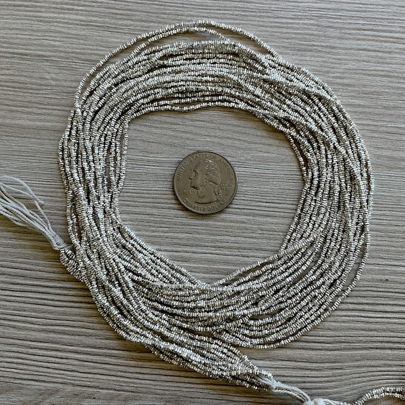 May include: Silver seed beads arranged in a circular pattern, with a quarter coin in the center for size reference. The beads are strung together, with a white thread visible at the bottom left.