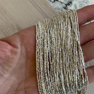 May include: Close-up of strands of silver-colored beads. The beads are small, square-shaped, and strung together, creating a shimmering effect. The beads are held in a hand, with a light-colored wooden surface in the background. The beads appear to be jewelry.