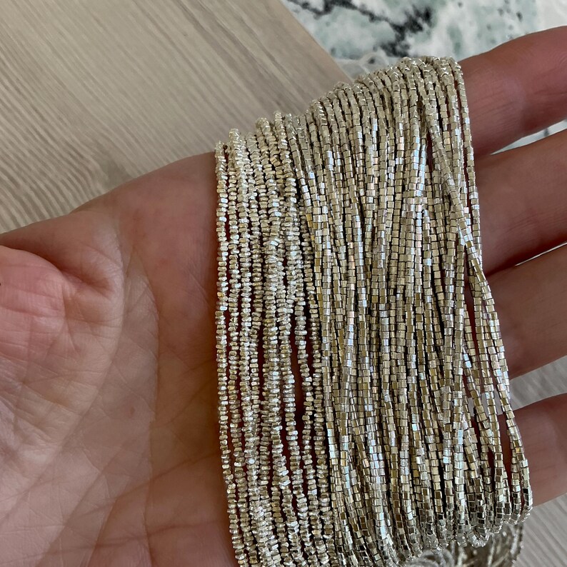 May include: Close-up of strands of silver-colored beads. The beads are small, square-shaped, and strung together, creating a shimmering effect. The beads are held in a hand, with a light-colored wooden surface in the background. The beads appear to be jewelry.