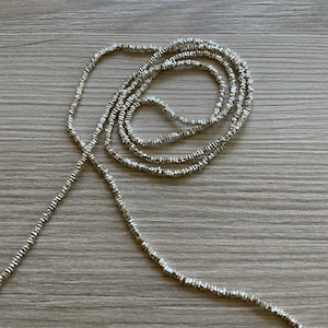 May include: A silver beaded necklace with a delicate, textured design. The necklace is draped across a wooden surface, showcasing its intricate details and reflective qualities. The beads appear to be small and evenly spaced.