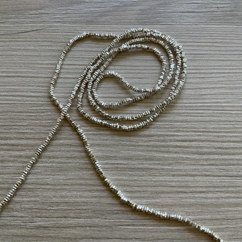 May include: A silver beaded necklace with a delicate, textured design. The necklace is draped across a wooden surface, showcasing its intricate details and reflective qualities. The beads appear to be small and evenly spaced.