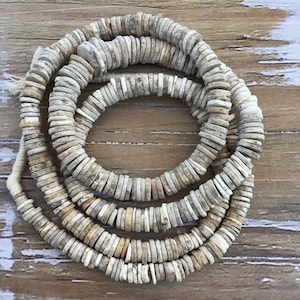 May include: A strand of white and beige shell beads, each bead is a flat disc shape. The beads are strung together on a white cord.
