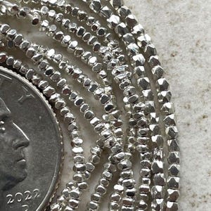 Può includere: Primo piano di una collana di perline in metallo argentato. Le perline sono piccole e hanno una forma irregolare sfaccettata. La collana è avvolta e sovrapposta, mostrando la texture delle perline.