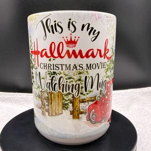 Könnte beinhalten: Weißer Keramikbecher mit dem Text "This is my Hallmark Christmas Movie Watching Mug" in Schwarz und Rot. Der Becher zeigt eine Winterszene mit einem roten LKW und einem Holzzaun.