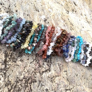 Könnte beinhalten: Eine Sammlung von Armbändern mit Edelstein-Chips in verschiedenen Farben, darunter Weiß, Grün, Lila, Schwarz, Gelb, Türkis, Rosa, Braun, Blau und Klar. Die Armbänder bestehen aus kleinen, unregelmäßig geformten Steinen, die aufgefädelt sind.