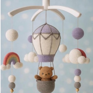 Op de afbeelding: Handgemaakte gehaakte baby mobile met een luchtballon, een regenboog, wolken en een teddybeer. De ballon is crème en lavendel, met een grijze mand voor de beer. Witte en grijze bollen hangen eronder.