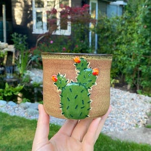 May include: A ceramic plant pot featuring a hand-painted cactus design. The pot is light brown with a darker brown rim. The cactus is green with small orange flowers and black details. The pot is held in a hand.