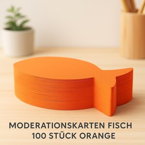 May include: A stack of bright orange fish-shaped cards. The cards are neatly stacked on a light-colored wooden surface. The text at the bottom reads "MODERATIONSKARTEN FISCH 100 STÜCK ORANGE". A small potted plant and a wooden pencil holder are in the background.