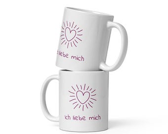 Può includere: Due tazze in ceramica bianca impilate. Ogni tazza presenta un disegno a forma di cuore rosa scuro con linee radianti. Il testo "Ich liebe mich" è stampato sotto il cuore su ogni tazza.