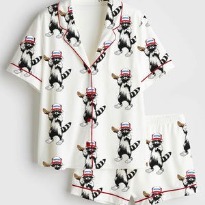 Op de afbeelding: Witte pyjamaset met een herhaald patroon van cartoon wasberen met rode hoeden en hotdogs. De top met korte mouwen en de shorts hebben een rode bies. Het woord "America" staat op de hoeden.