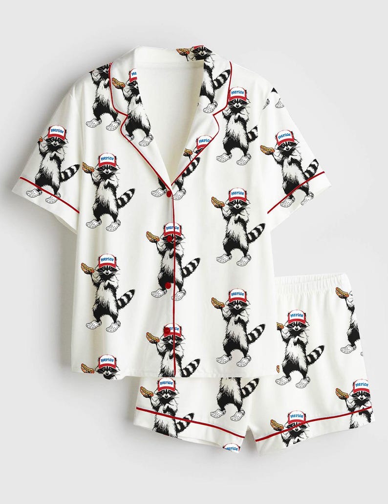 Op de afbeelding: Witte pyjamaset met een herhaald patroon van cartoon wasberen met rode hoeden en hotdogs. De top met korte mouwen en de shorts hebben een rode bies. Het woord "America" staat op de hoeden.