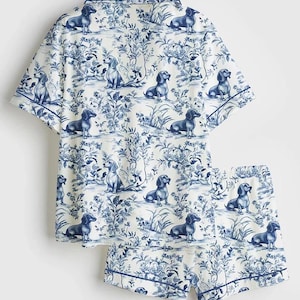 Puede incluir: Un conjunto de pijama blanco con un estampado de toile de Jouy azul con perros salchicha y elementos florales. El conjunto incluye una camisa de manga corta y un pantal&oacute;n corto, ambos con un borde azul.