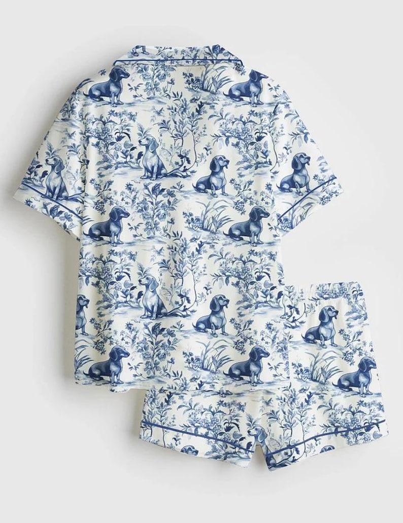 Puede incluir: Un conjunto de pijama blanco con un estampado de toile de Jouy azul con perros salchicha y elementos florales. El conjunto incluye una camisa de manga corta y un pantal&oacute;n corto, ambos con un borde azul.