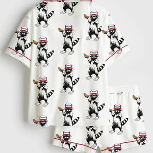 Op de afbeelding: Witte pyjamaset met een herhalend patroon van cartoon wasberen. Elke wasbeer draagt een rood, wit en blauw hoedje en houdt een gebakje vast. De set bevat een shirt met korte mouwen en bijpassende shorts, beide afgezet met rode biezen.