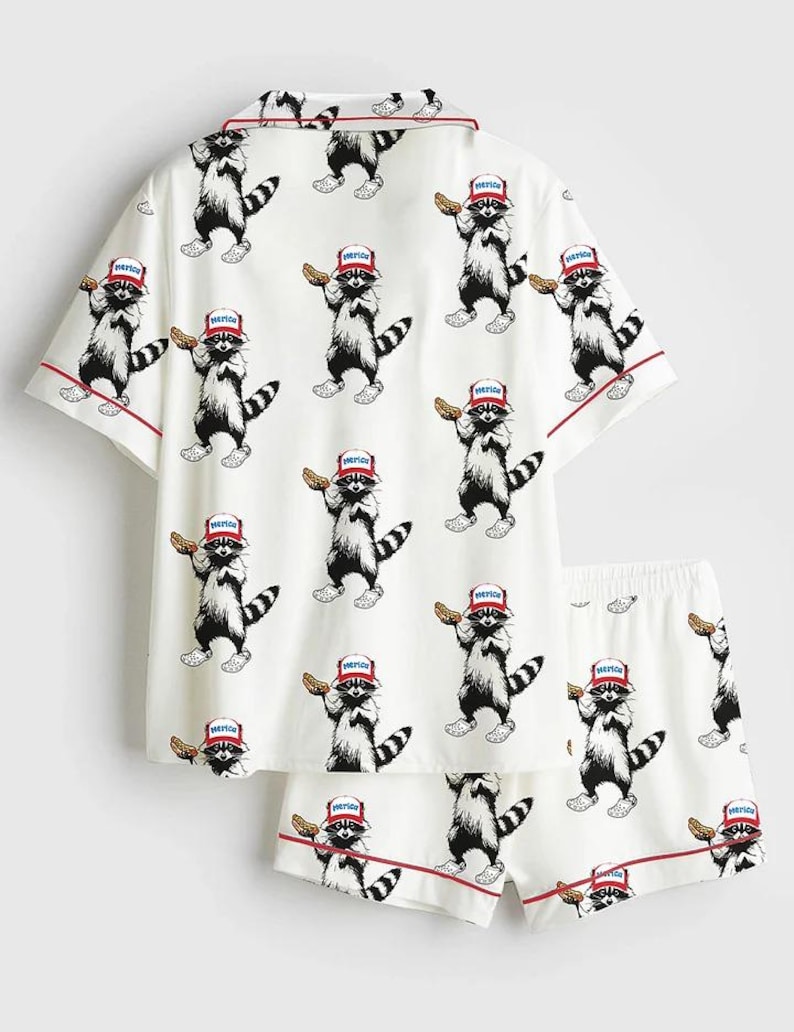 Op de afbeelding: Witte pyjamaset met een herhalend patroon van cartoon wasberen. Elke wasbeer draagt een rood, wit en blauw hoedje en houdt een gebakje vast. De set bevat een shirt met korte mouwen en bijpassende shorts, beide afgezet met rode biezen.