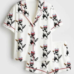 Op de afbeelding: Witte pyjamaset met een herhalend patroon van wasberen met rode hoeden en eten. De top met korte mouwen en de shorts hebben rode biezen. De wasberen zijn zwart-wit met het woord "America" op hun hoeden.