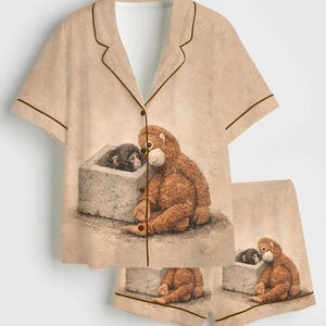 May include: Tan pajama set featuring a short-sleeved button-down shirt and matching shorts. The set is adorned with a whimsical illustration of two monkeys. The shirt and shorts have brown trim.