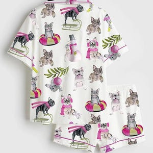 May include: White pajama set featuring a repeating pattern of French bulldogs in winter scenes. The short-sleeved top and shorts have pink trim and are decorated with dogs wearing scarves, hats, and riding sleds. The set includes a snowman.
