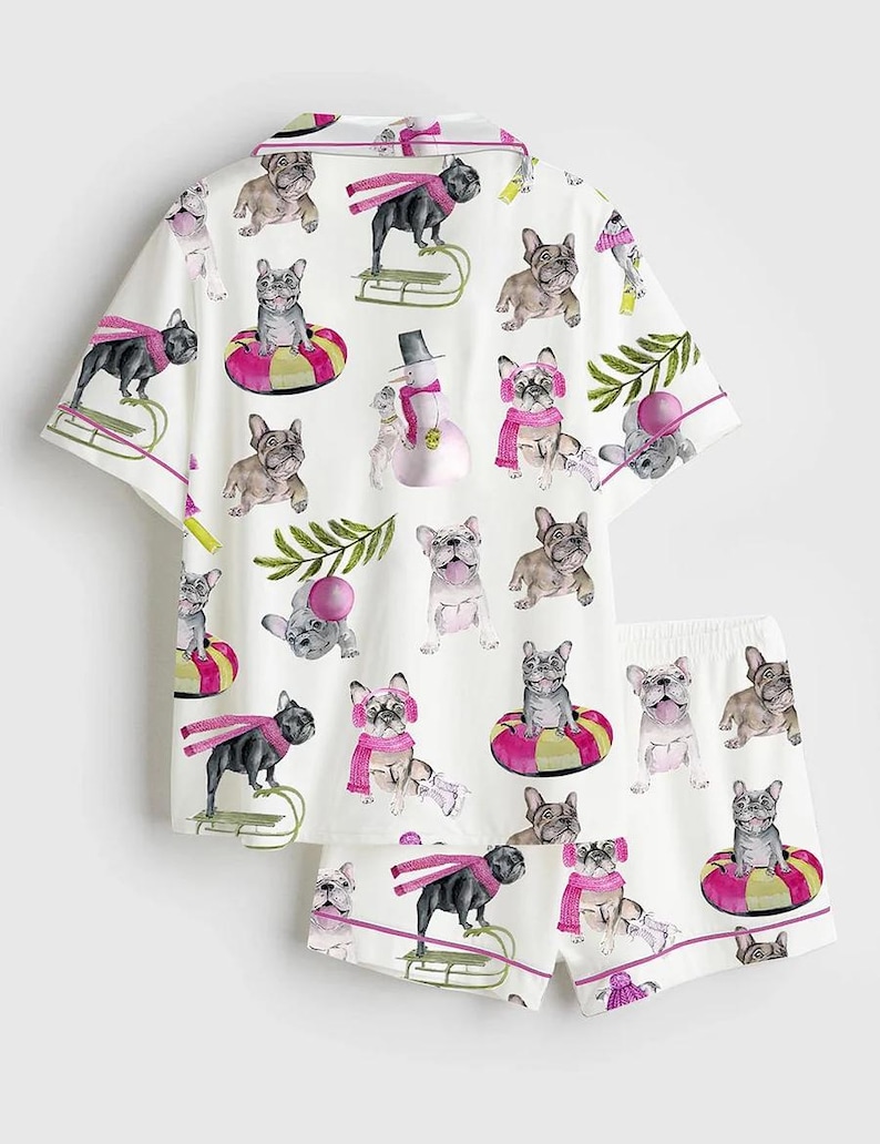 May include: White pajama set featuring a repeating pattern of French bulldogs in winter scenes. The short-sleeved top and shorts have pink trim and are decorated with dogs wearing scarves, hats, and riding sleds. The set includes a snowman.