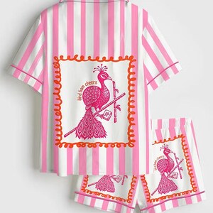 May include: Pink and white striped pajama set. The short-sleeved top and shorts feature a pink peacock graphic with the text "bird bam cheers" in a decorative frame. The set has pink trim.