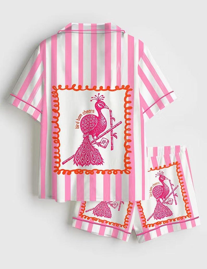 May include: Pink and white striped pajama set. The short-sleeved top and shorts feature a pink peacock graphic with the text "bird bam cheers" in a decorative frame. The set has pink trim.